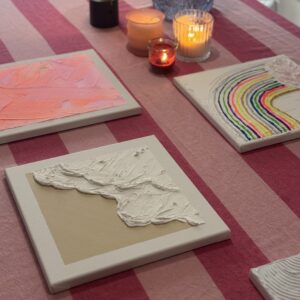 Drei fertige Leinwände in Rosa, mit Regenbogenmotiv und mit weißer Struktur, daneben Kerzen und eine orange Lampe