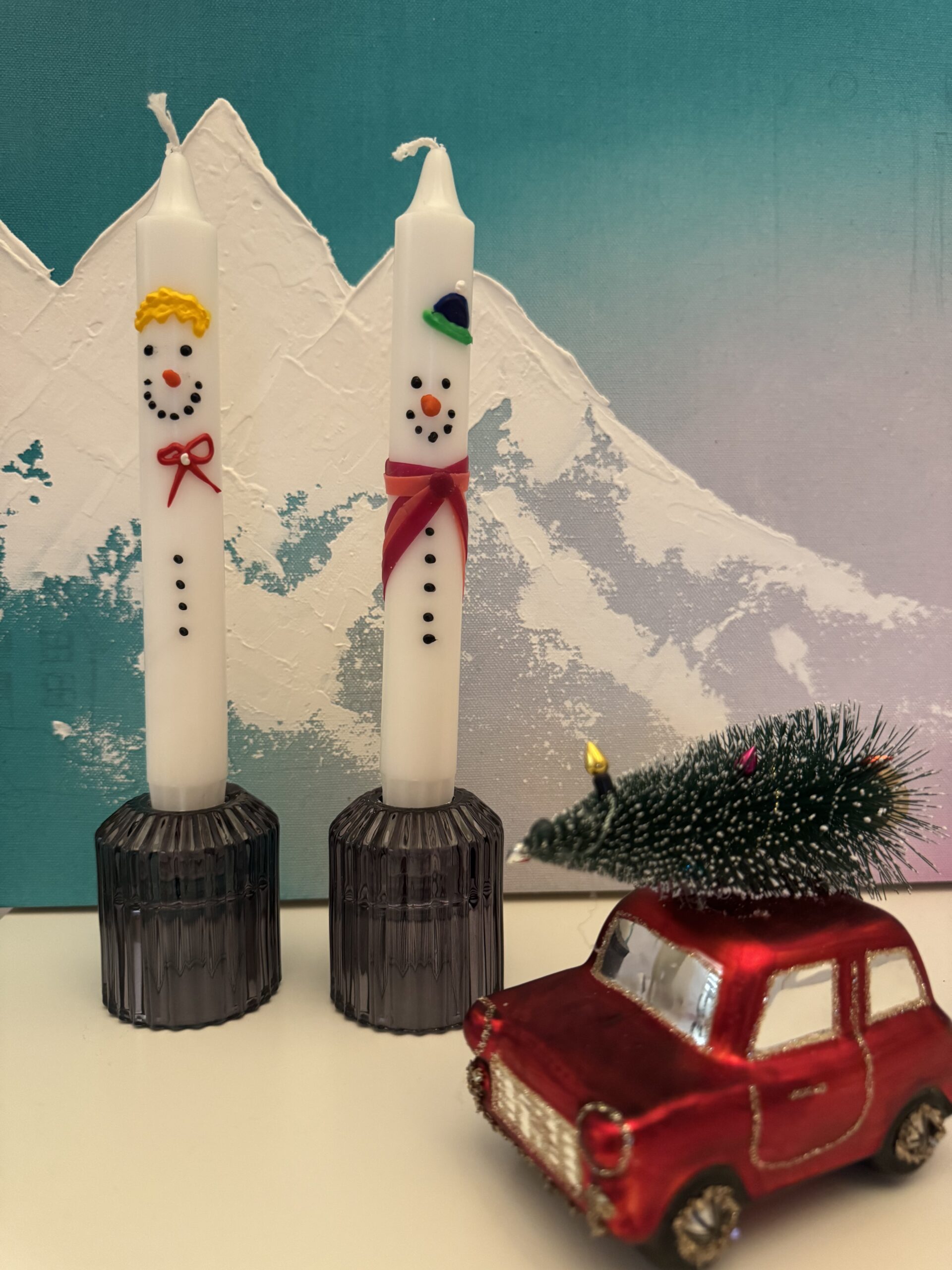Bemalte weiße Kerzen mit winterlichen Motiven, daneben ein kleines rotes Spielzeugauto als Dekoration