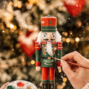Hände bemalen einen Holz-Nussknacker in Rot und Grün mit weißem Bart, daneben eine Farbpalette, im Hintergrund ein leuchtender Weihnachtsbaum